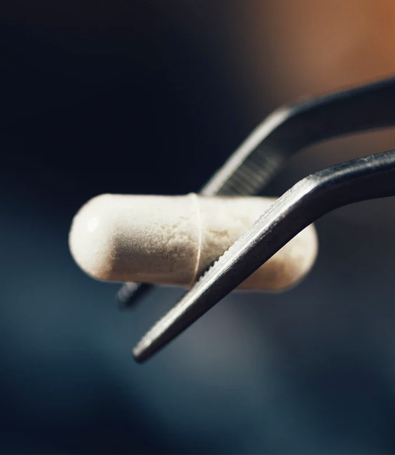 A close-up of a white capsule being held by metal tweezers against a blurred dark background.