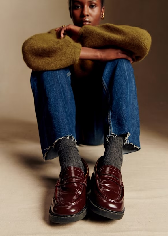 Person seated with arms resting on knees, wearing a greenish-brown sweater, blue jeans, gray socks, and burgundy penny loafers, photographed against a neutral background.