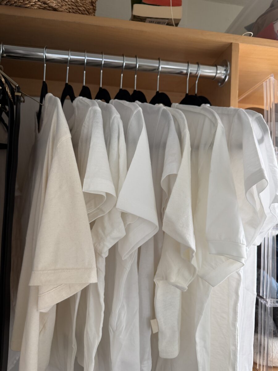 Several white T-shirts hang neatly on black hangers in a wooden closet, with sunlight shining on the clothing.