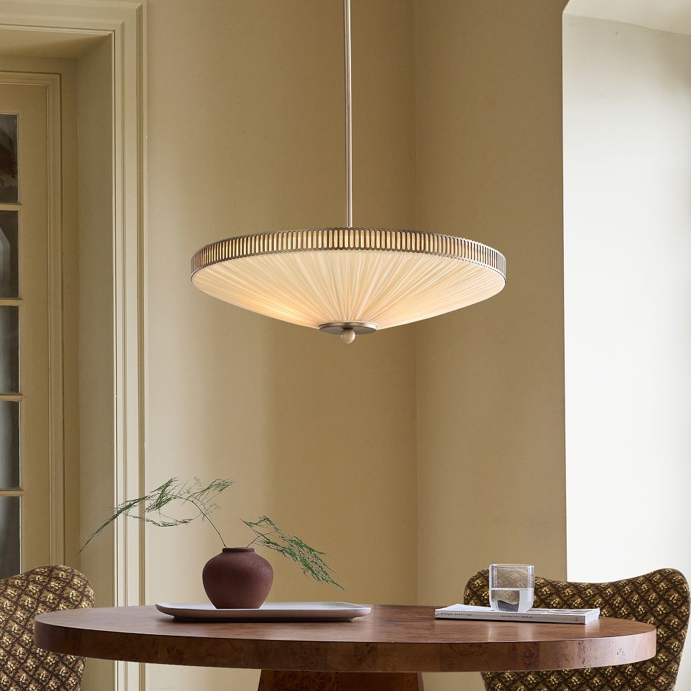 A round pendant light hangs above a small circular table with a brown vase, a branch, a white plate, and a glass of water in a neutral-toned room.