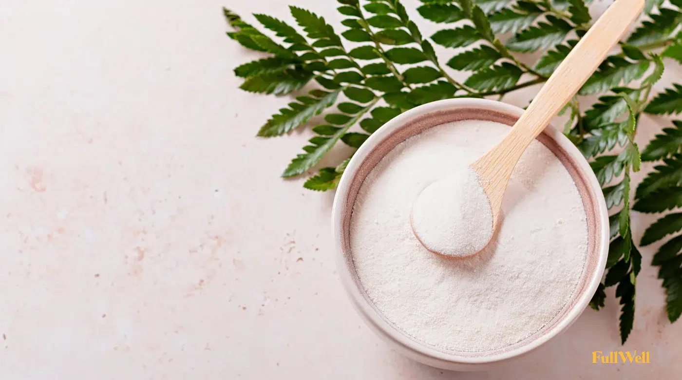 A bowl of white powder with a wooden spoon rests on a light surface, next to green leaves.
