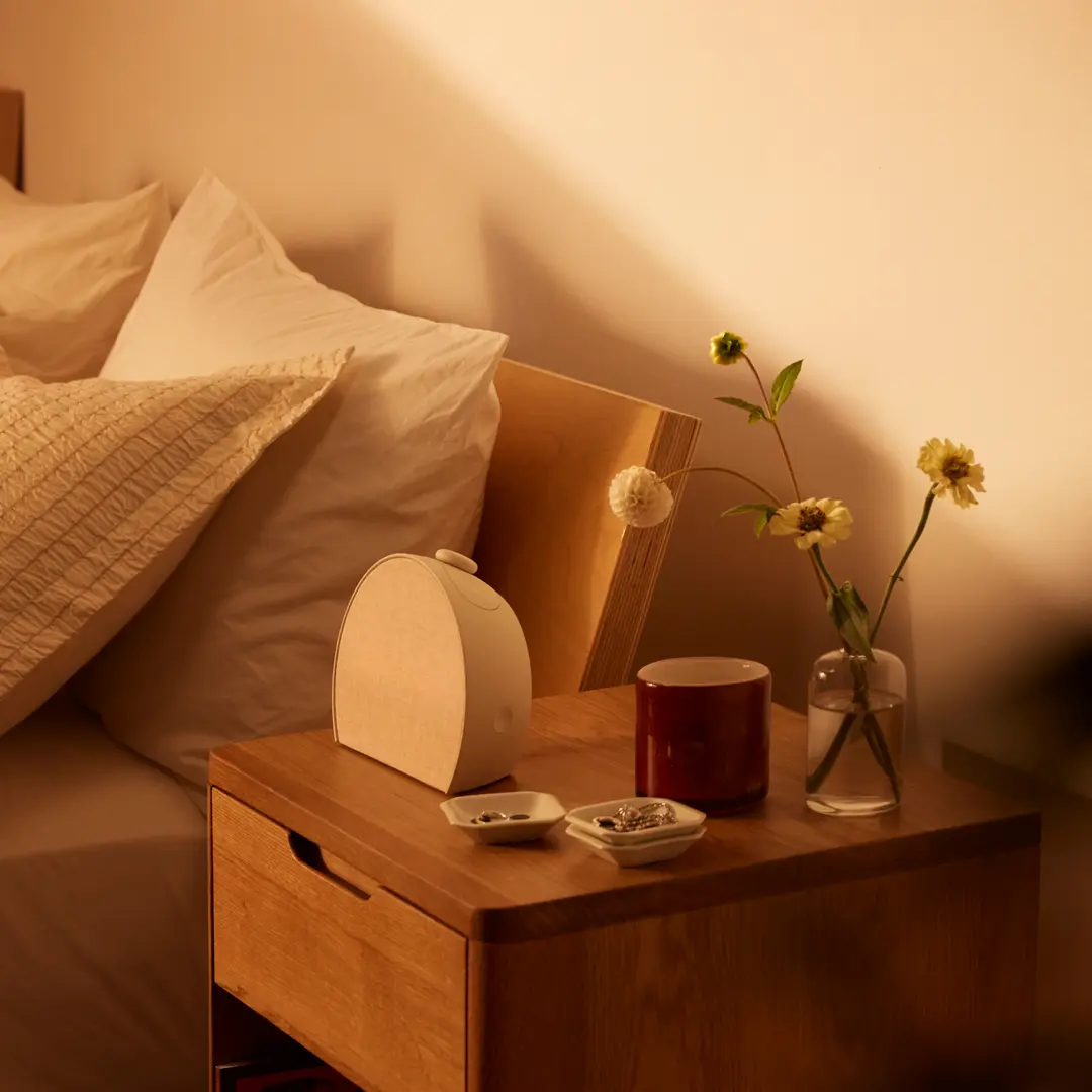A wooden bedside table with a red mug, a glass vase with yellow flowers, two small dishes, and a white device beside a bed with white pillows.