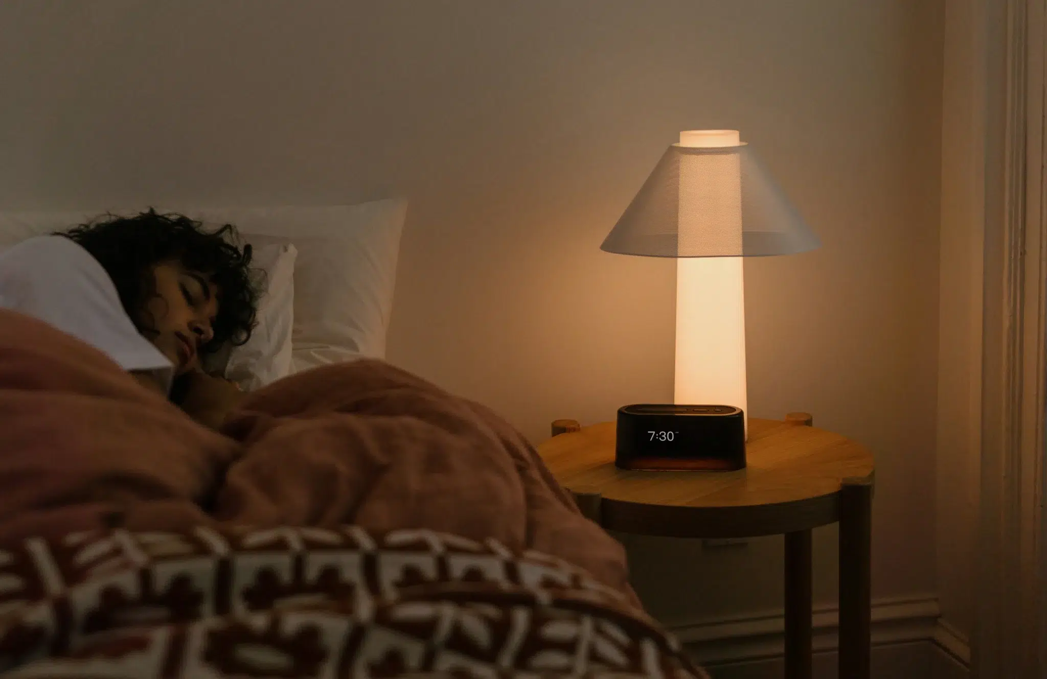 A person sleeps in bed beside a wooden nightstand with a lit lamp and a digital clock displaying 7:30.