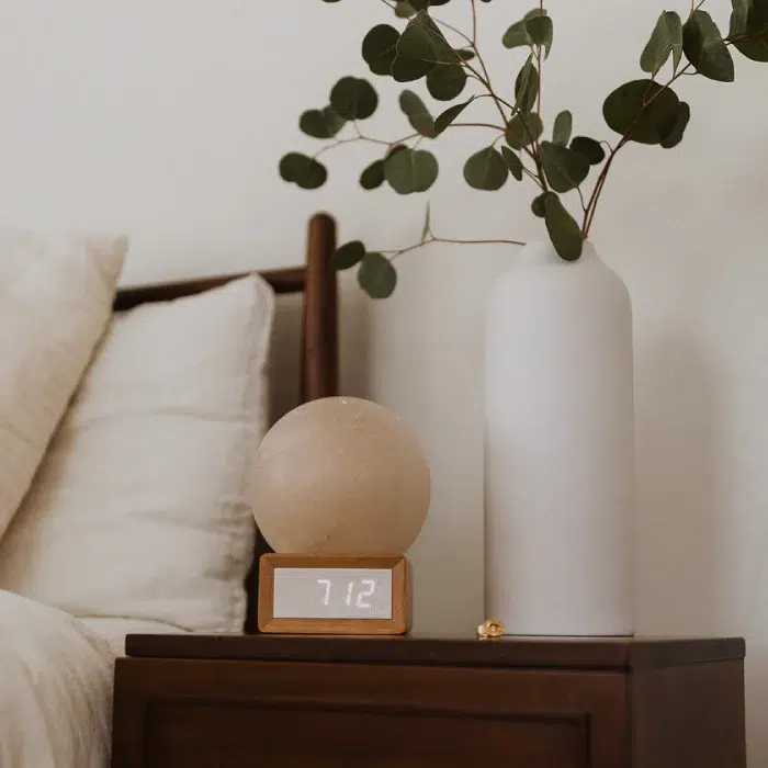 A wooden nightstand with a digital clock displaying 7:12, a round lamp, a tall white vase with green branches, and part of a bed with white bedding.