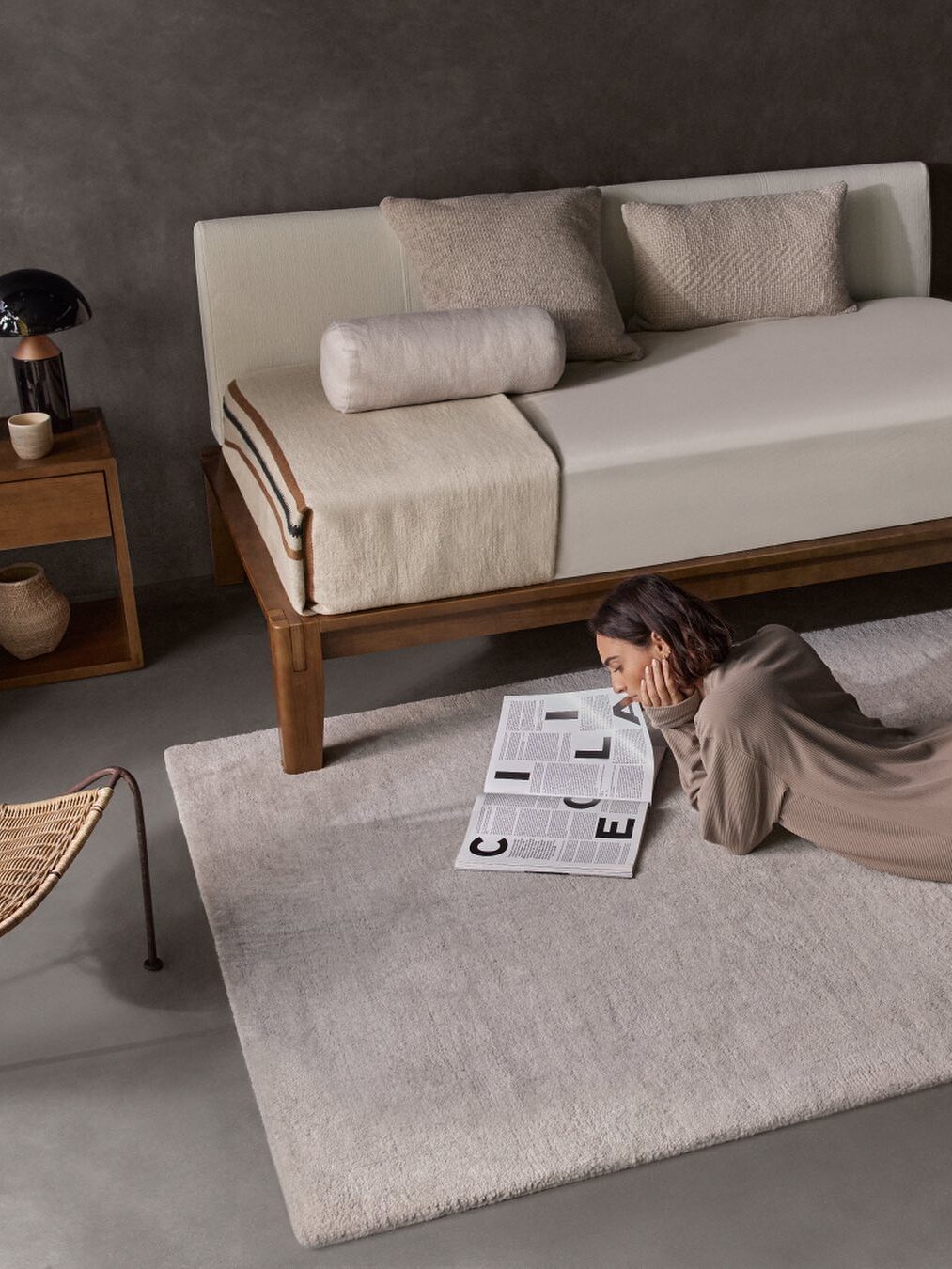 A woman lies on a light rug reading a newspaper in a minimalist room with a wooden daybed, cushions, a side table, and a rattan chair.