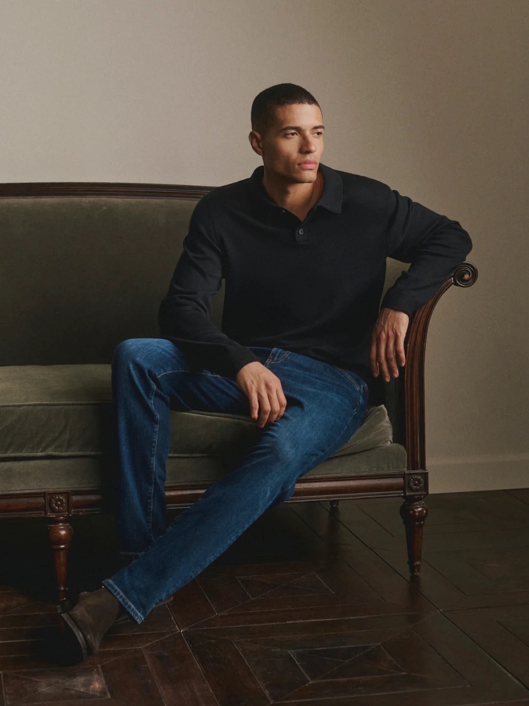 A person in a dark long-sleeve shirt and blue jeans sits on a green velvet sofa, looking to the side in a neutral-toned room.