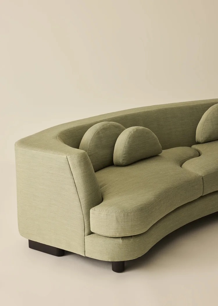A curved, light green sofa with matching rounded cushions and dark wooden legs, set against a plain beige background.
