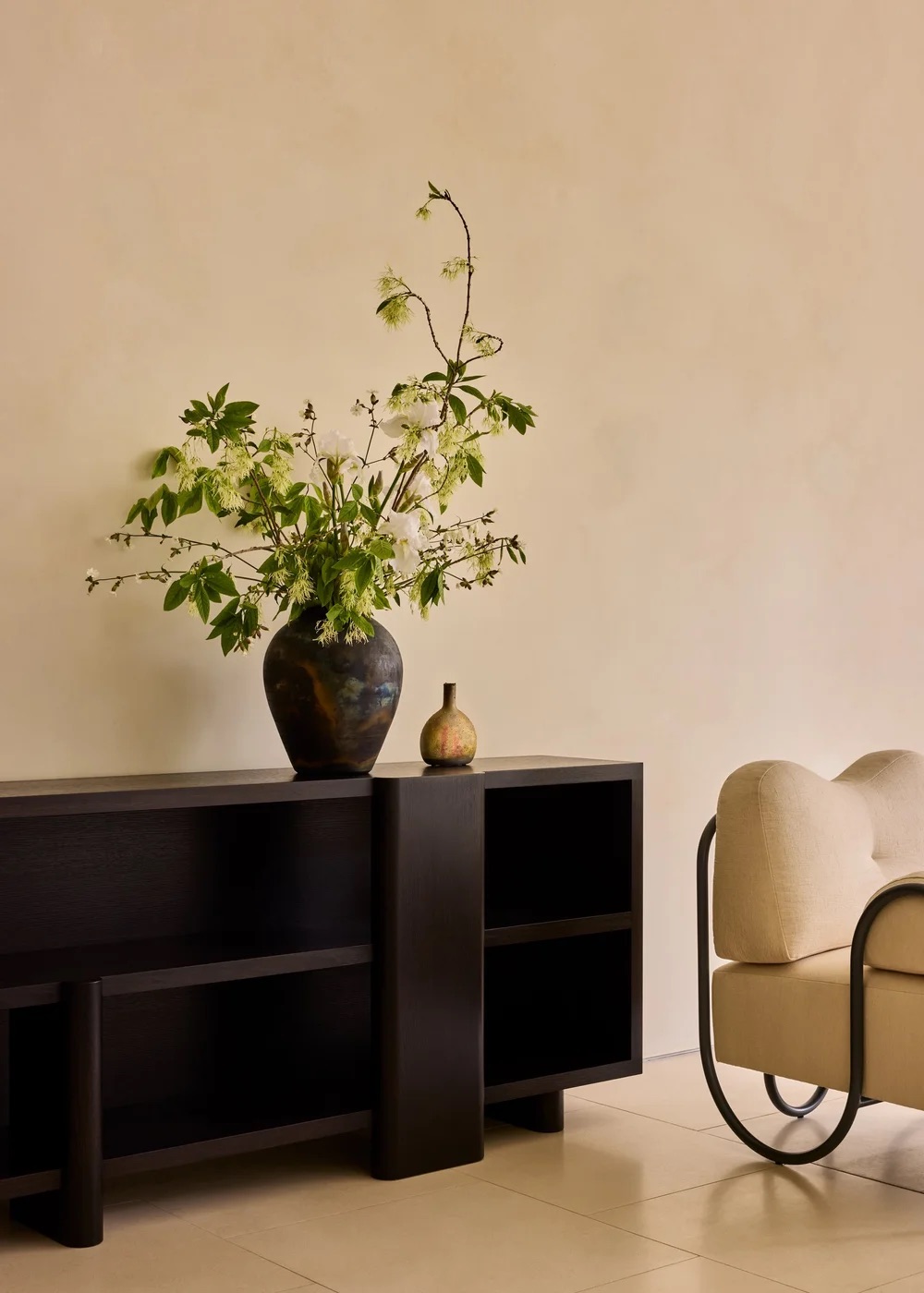 A dark wooden console holds a large vase with green and white flowers and a small brown vessel, next to a cream-colored armchair with black metal arms against a beige wall.