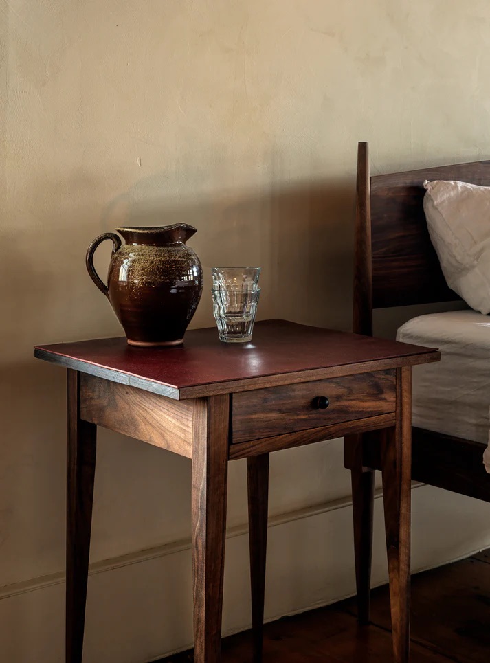 A brown ceramic pitcher and a clear glass sit on a wooden bedside table next to a bed with white bedding against a beige wall.