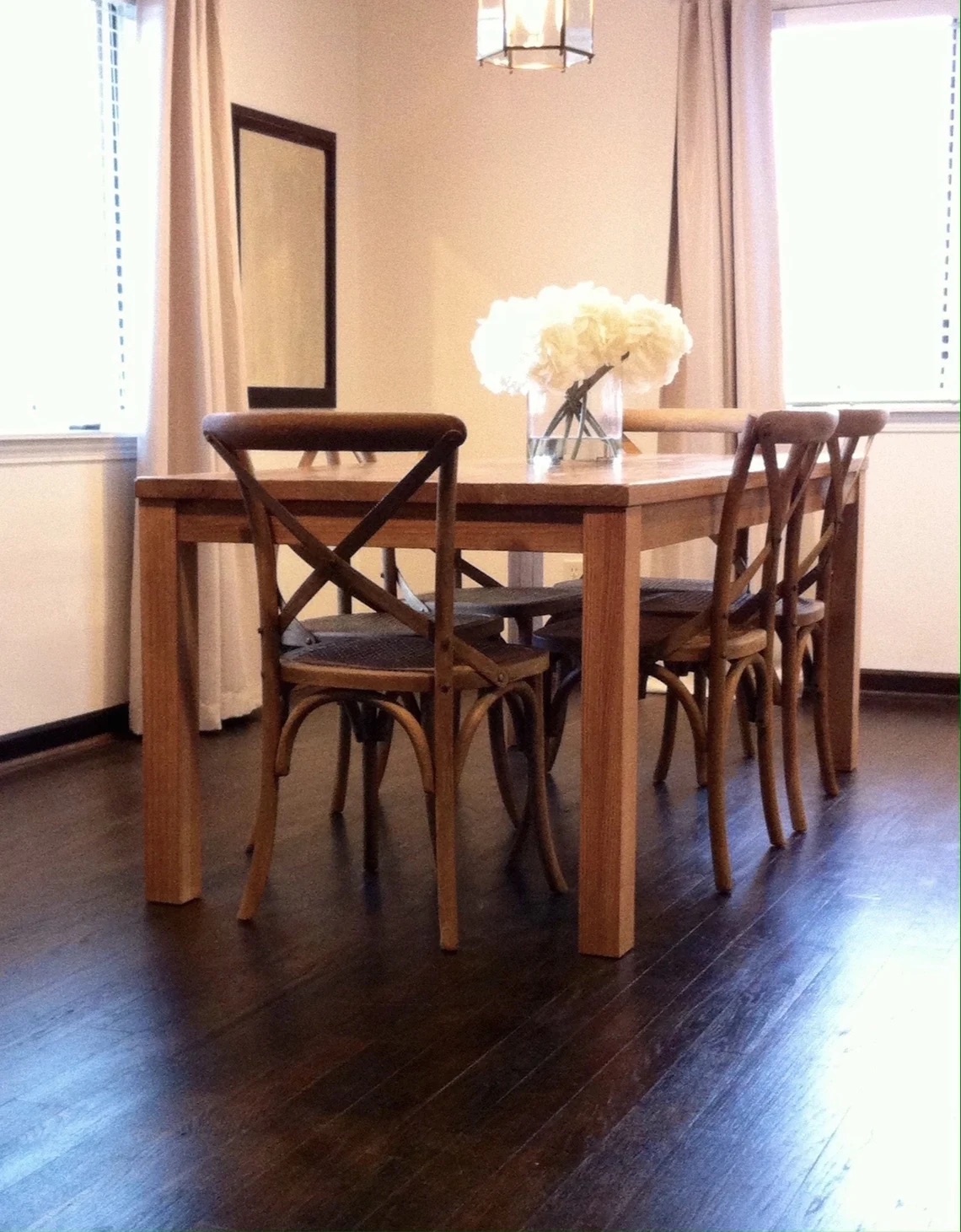 A wooden dining table with four chairs and a glass vase of white flowers on top, set in a room with dark wood floors and large windows with beige curtains.