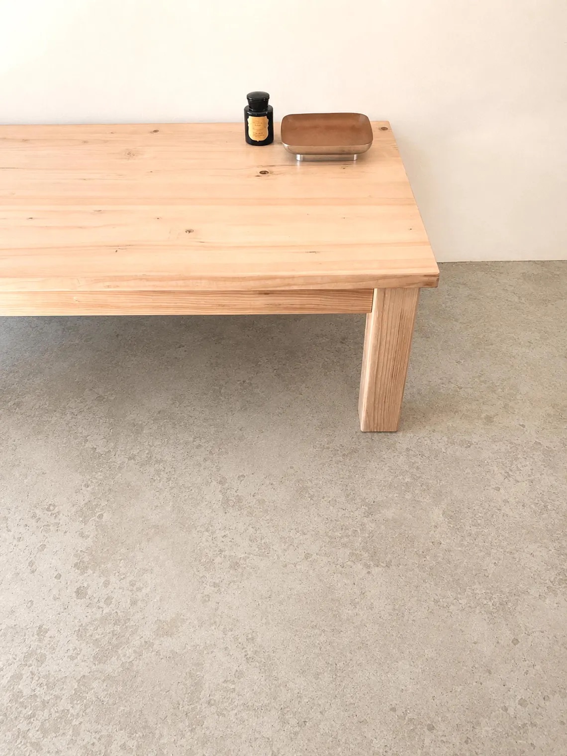 A light wooden table with square legs holds a small black jar, a candle, and a rectangular tray, set against a plain beige wall and a concrete floor.