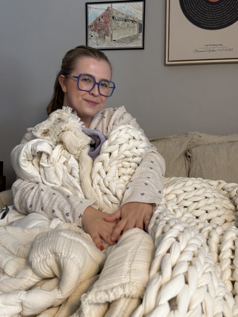 A person in glasses and pajamas sits on a couch wrapped in thick, cream-colored knit blankets. Framed artwork hangs on the wall behind them.
