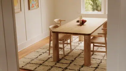 A wooden dining table with three chairs sits on a patterned rug in a bright room with light walls, artwork, and a window showing greenery outside.