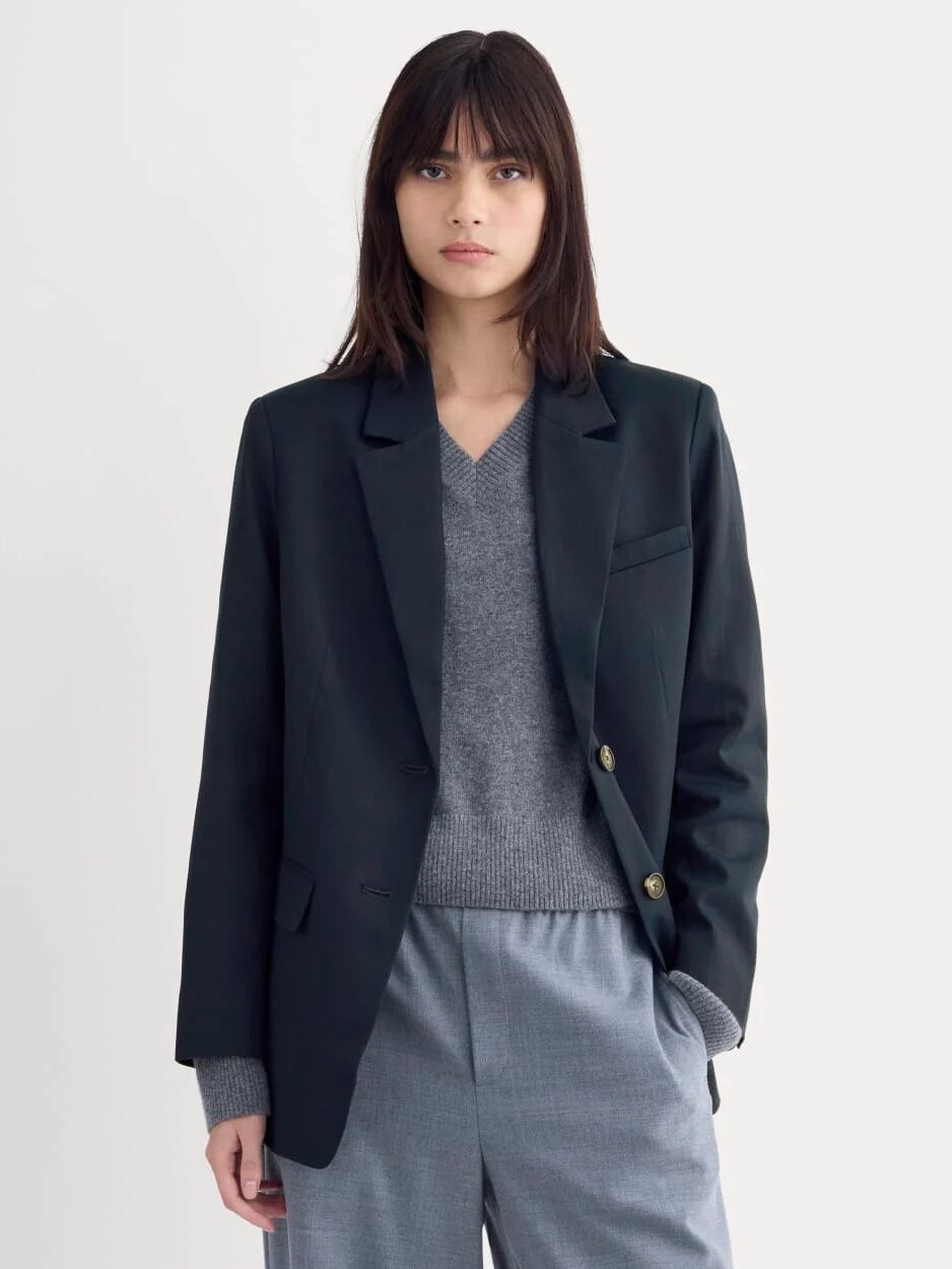 A woman with straight brown hair wears a dark blazer over a grey v-neck sweater and light grey trousers, standing against a plain light background.