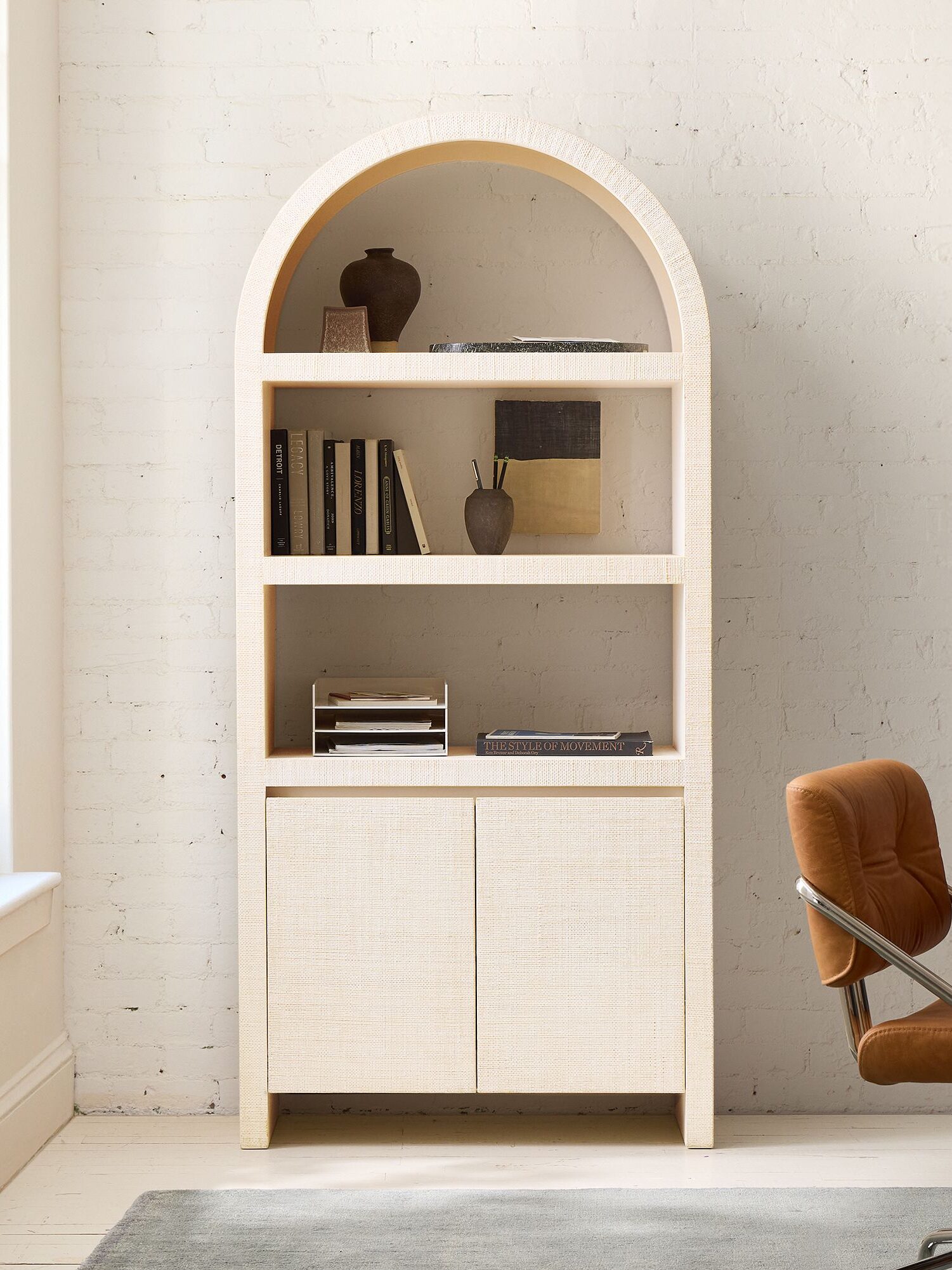 A white arched bookshelf with three open shelves holding books and decor items, above a two-door cabinet, stands in a bright room next to a window and desk.