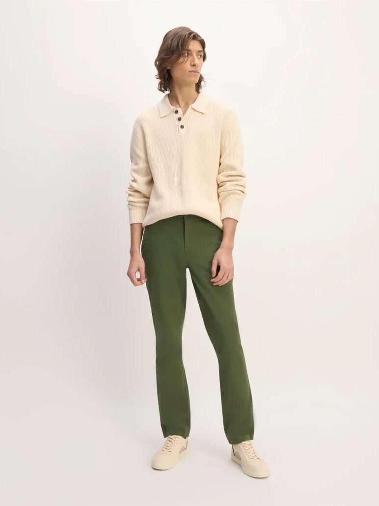 A person wearing a cream-colored sweater, green pants, and beige shoes stands against a plain light background, looking to the side.