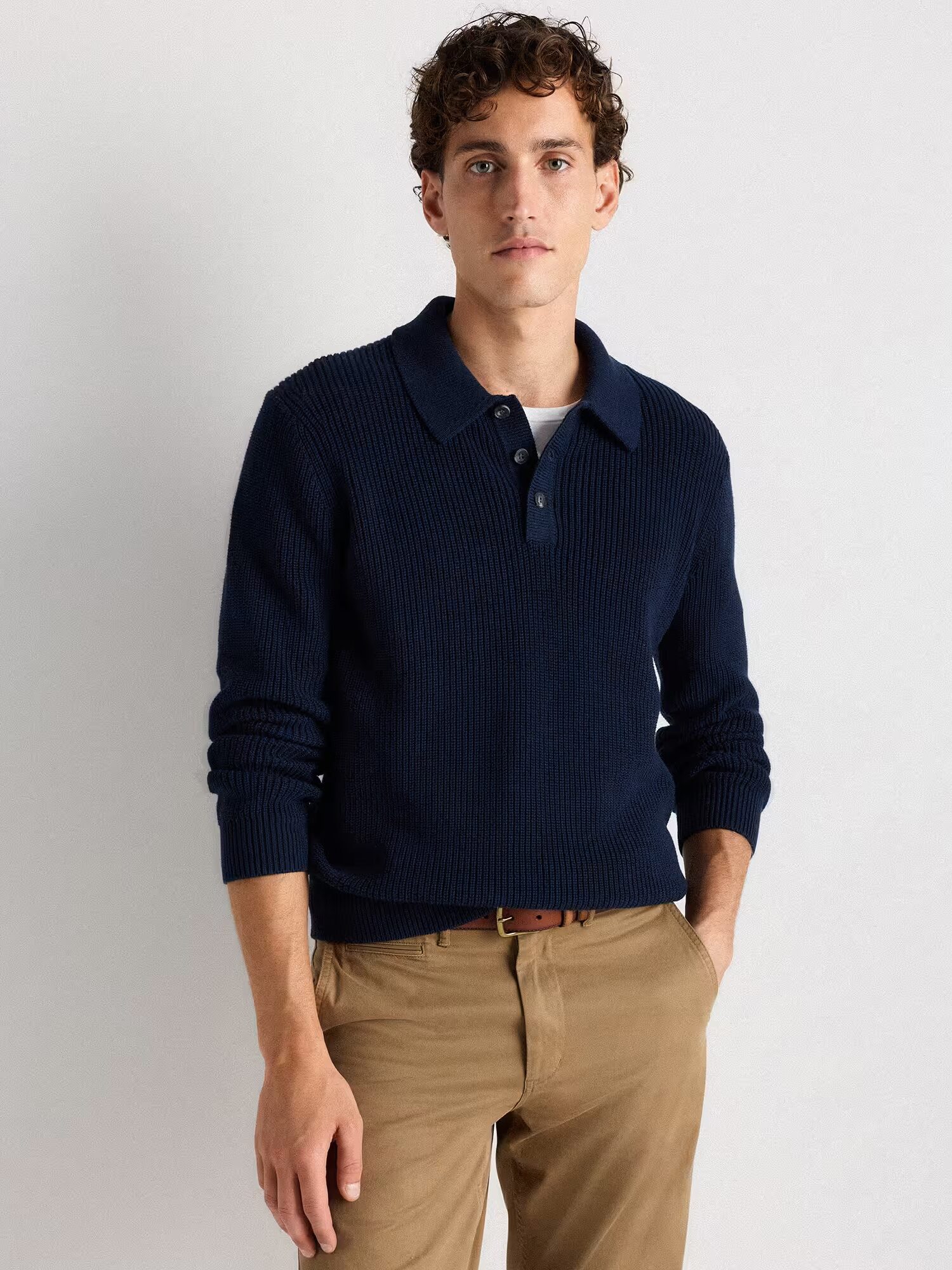 A man wearing a navy blue knit collared sweater over a white shirt and khaki pants stands against a plain light background.