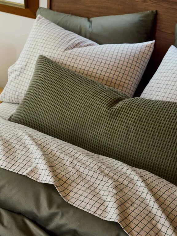 A neatly made bed with a green textured pillow, green bedding, and white grid-patterned pillowcases, next to a wooden nightstand with a yellow lamp.