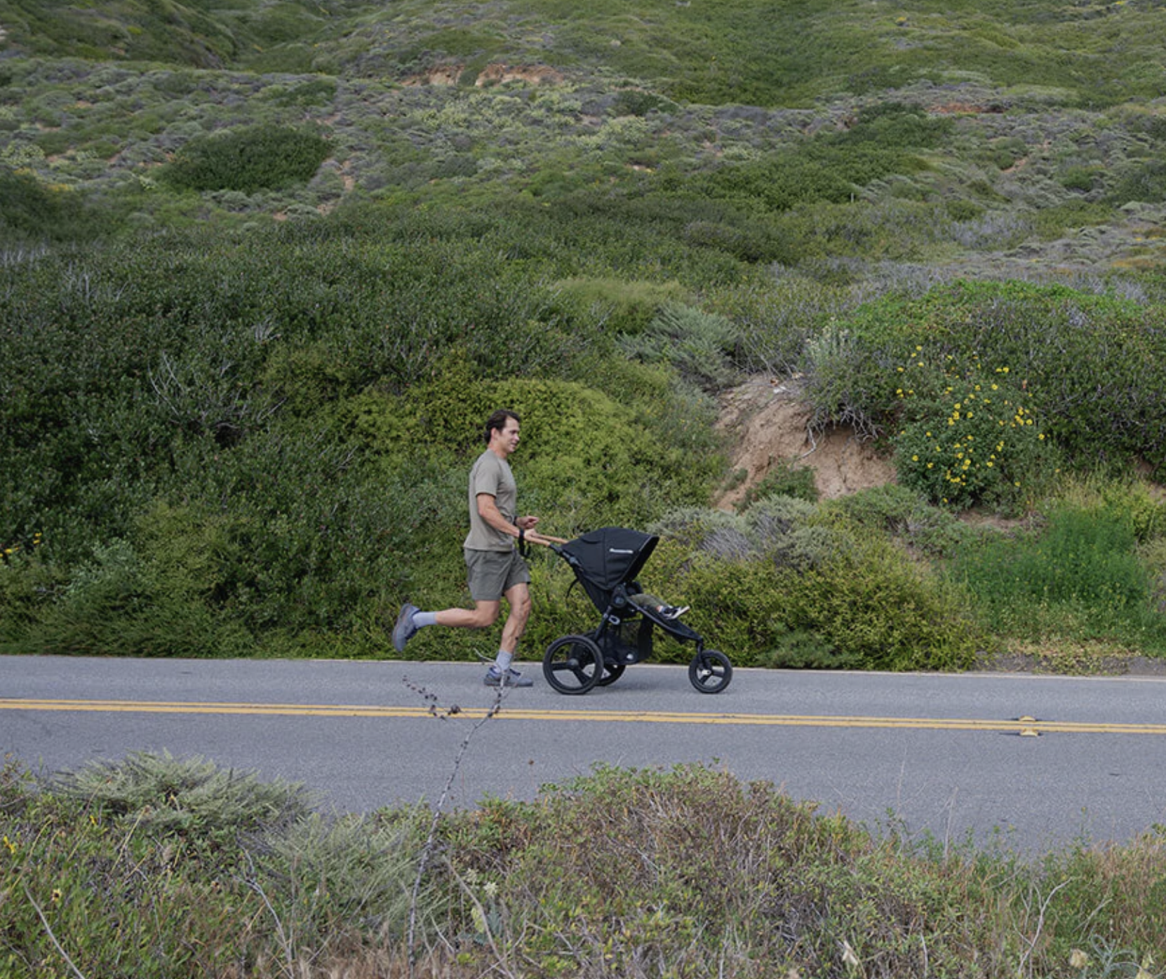 A man jogs on a paved road while pushing a black stroller, surrounded by green hills and vegetation.