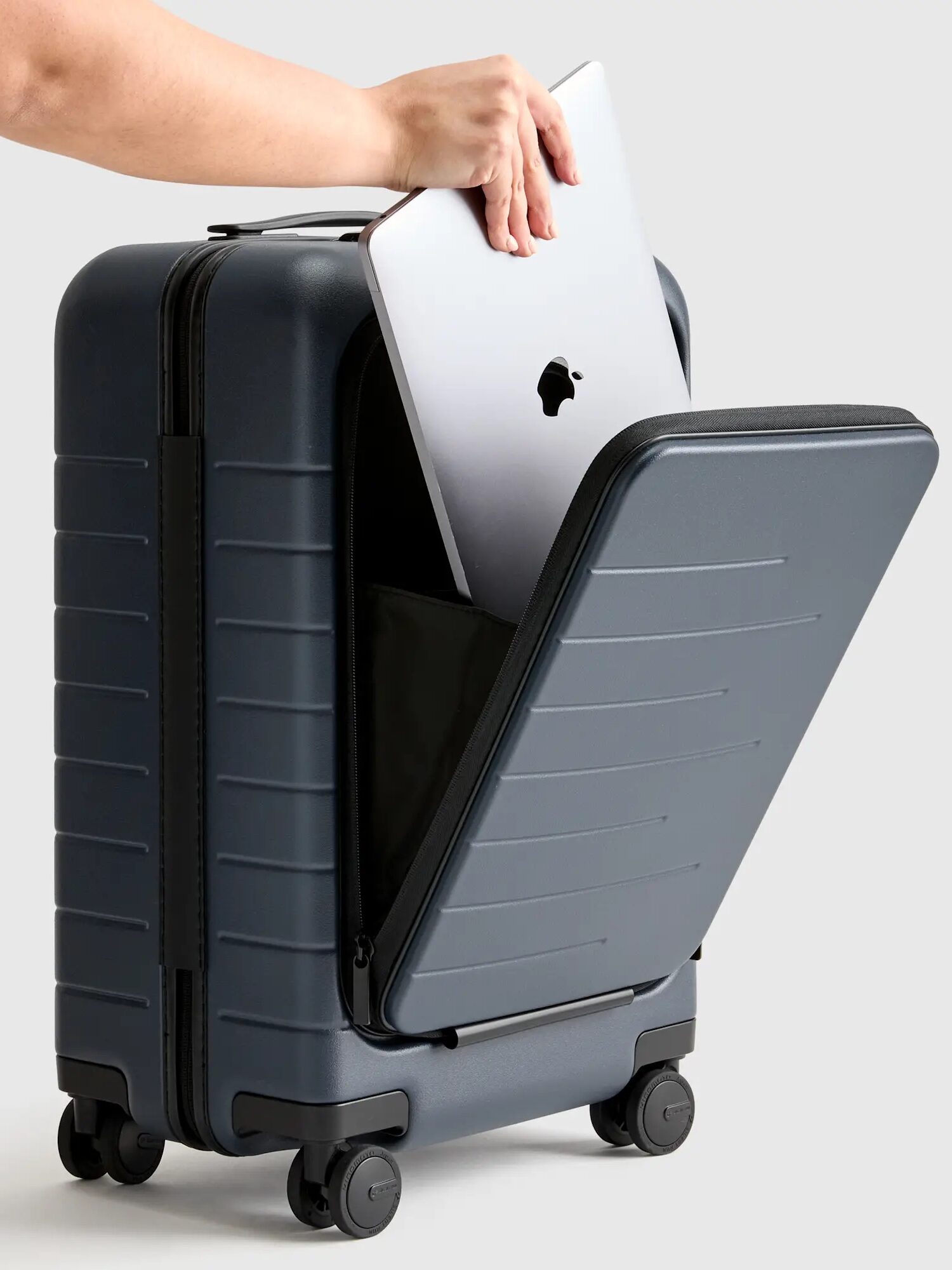 A hand places a silver laptop into the front pocket of a dark gray hard-shell carry-on suitcase with four wheels.