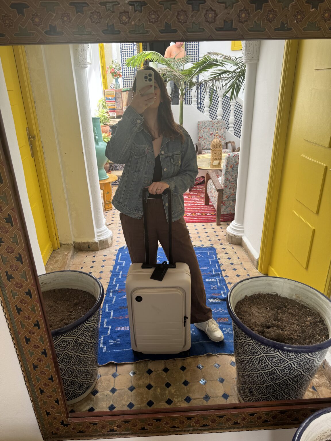 A person stands in front of a mirror holding a phone and suitcase, wearing a denim jacket and brown pants, in a brightly decorated room with yellow doors and patterned pots.
