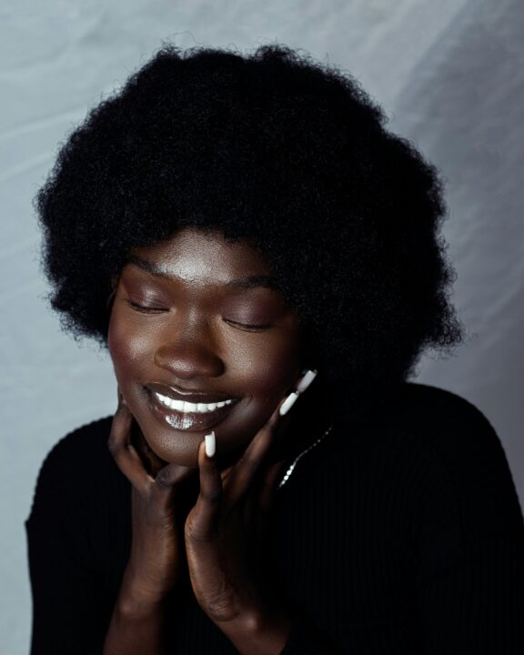 A person with dark skin and an afro hairstyle smiles with closed eyes, gently resting their hands on their face, against a light gray background.