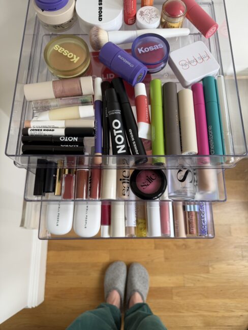 Two clear drawers filled with neatly organized makeup products, including lipsticks, mascaras, and blushes; a pair of feet in gray slippers visible on a wooden floor below.