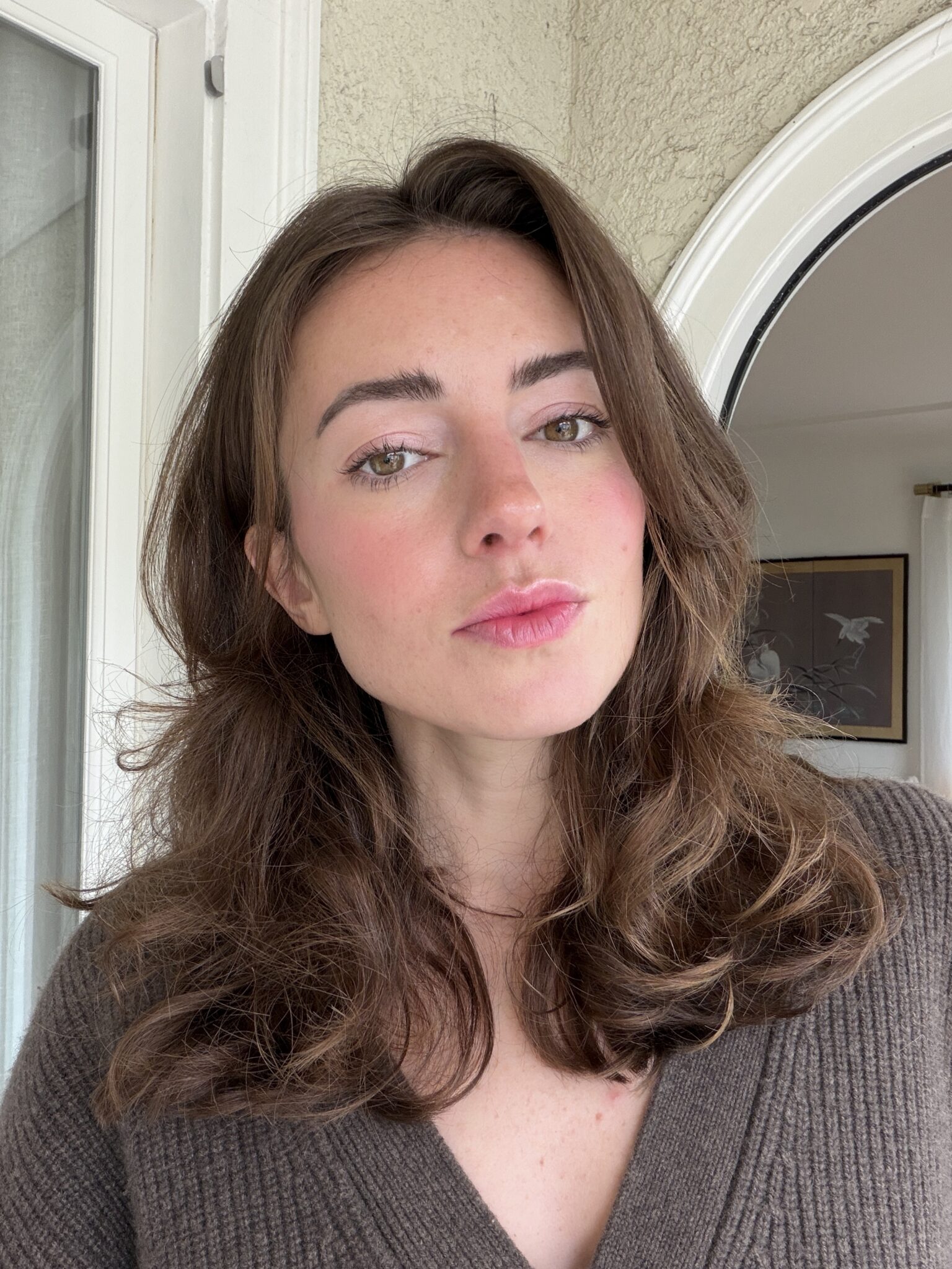 A woman with long brown hair and a neutral expression stands indoors near a door, wearing a brown V-neck sweater.