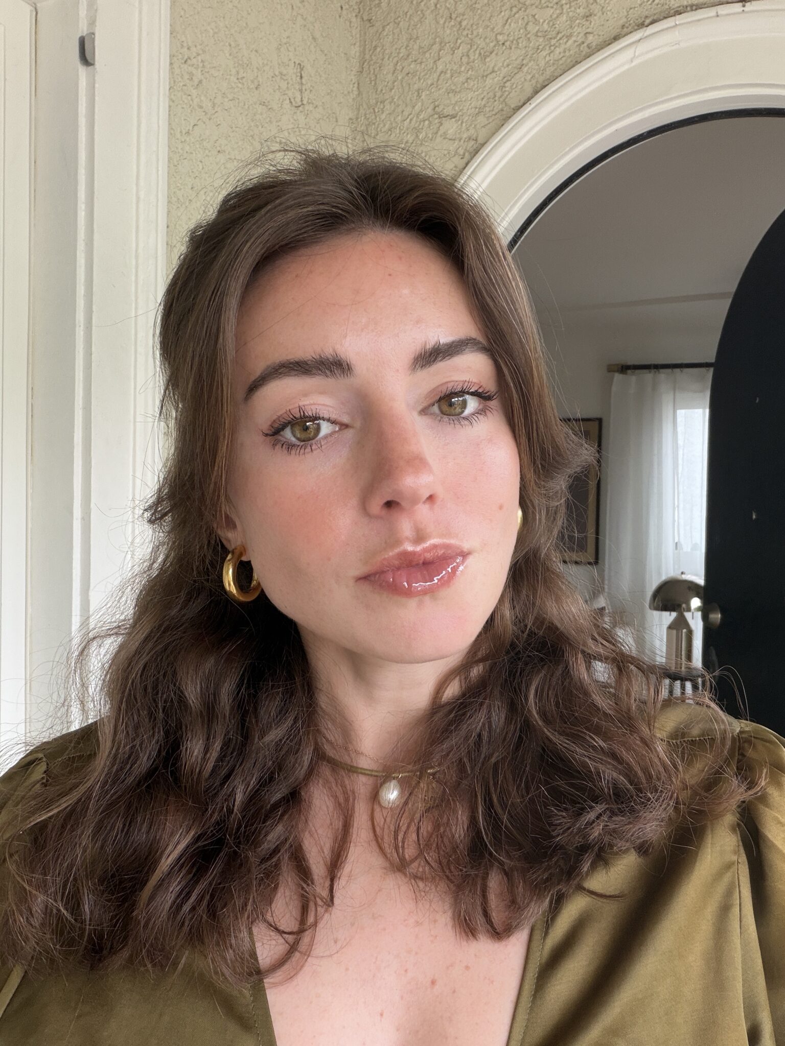 A woman with wavy brown hair, wearing a green top, gold hoop earrings, and a pearl necklace, poses indoors in front of a textured wall and a partially visible door.