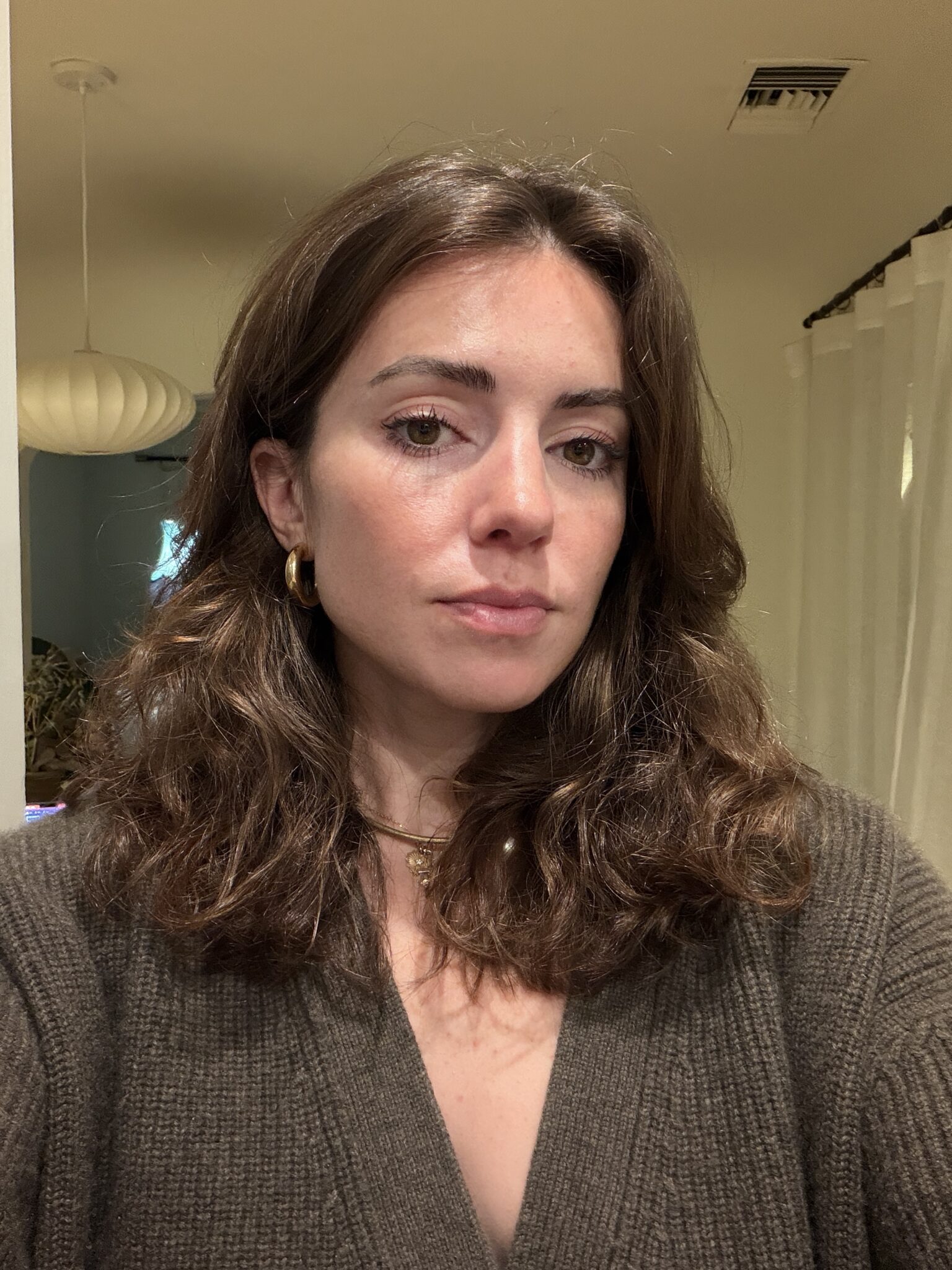 Woman with wavy brown hair wearing gold hoop earrings and a brown sweater looks at the camera indoors, with neutral expression and soft lighting.