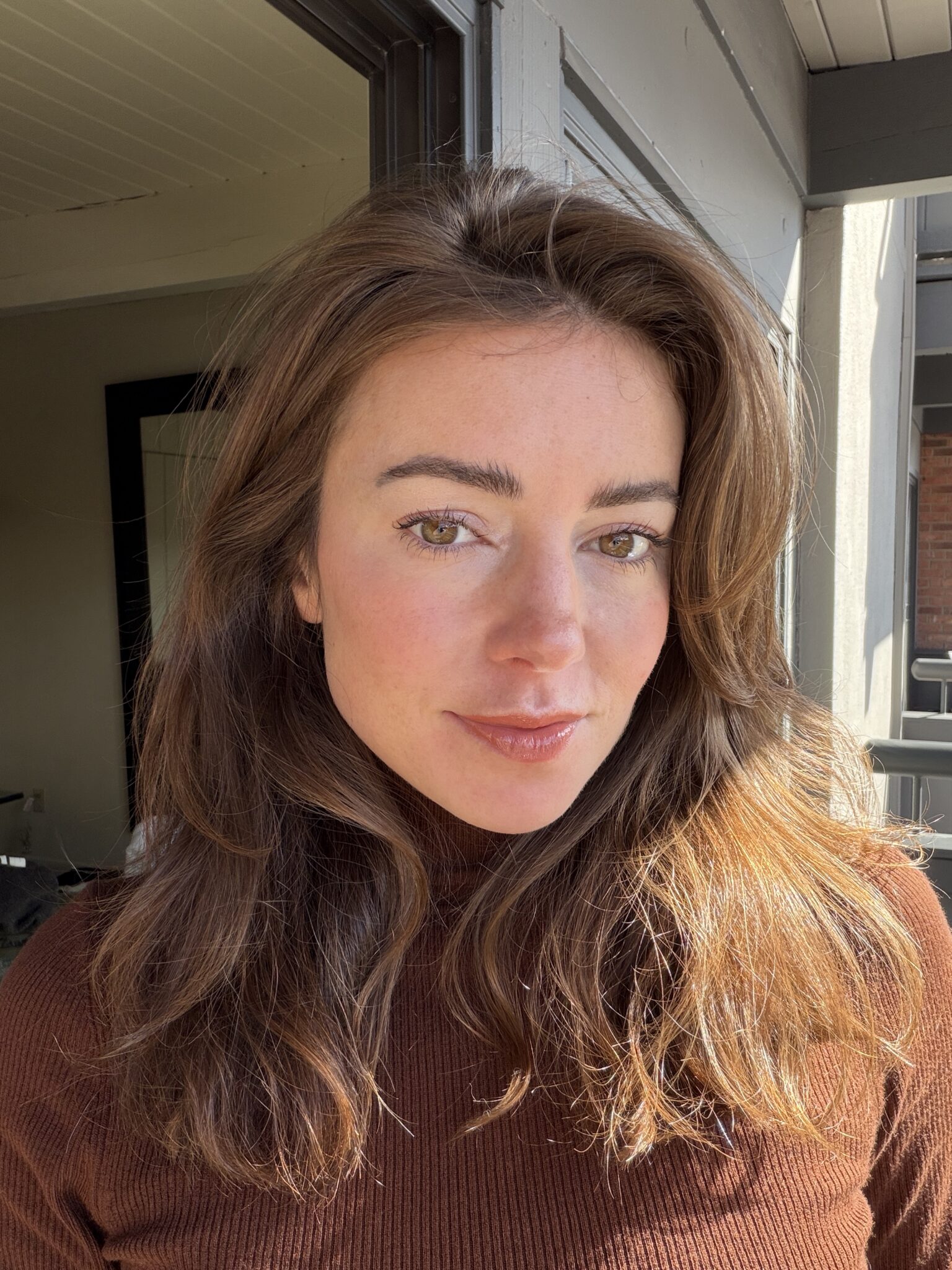 A woman with long brown hair and a brown turtleneck sweater stands indoors near a window, looking directly at the camera.