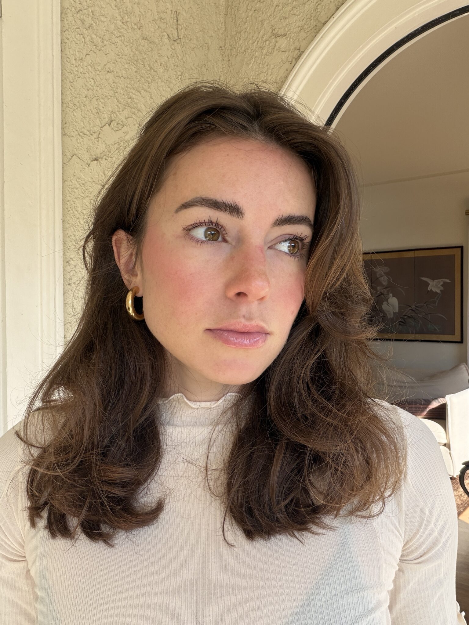 A woman with wavy brown hair and gold hoop earrings stands indoors, wearing a light-colored top, looking slightly to the side.