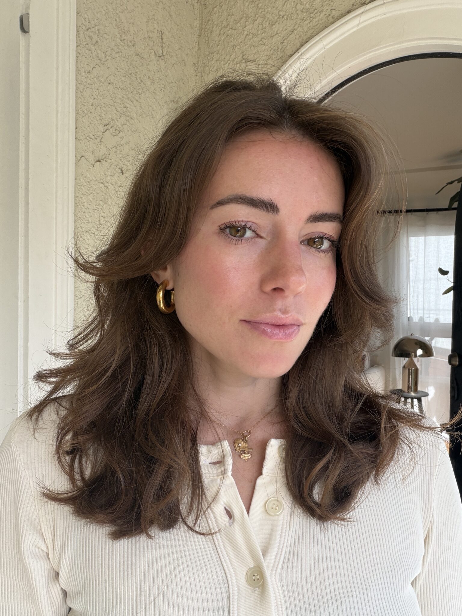 A woman with wavy brown hair and gold hoop earrings stands indoors, wearing a cream-colored buttoned top and a necklace, looking at the camera.