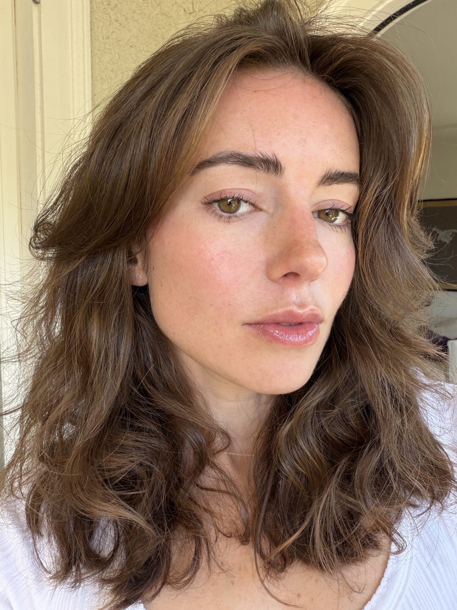 A person with wavy brown hair and light green eyes is looking at the camera, wearing a white top, in a well-lit indoor setting.