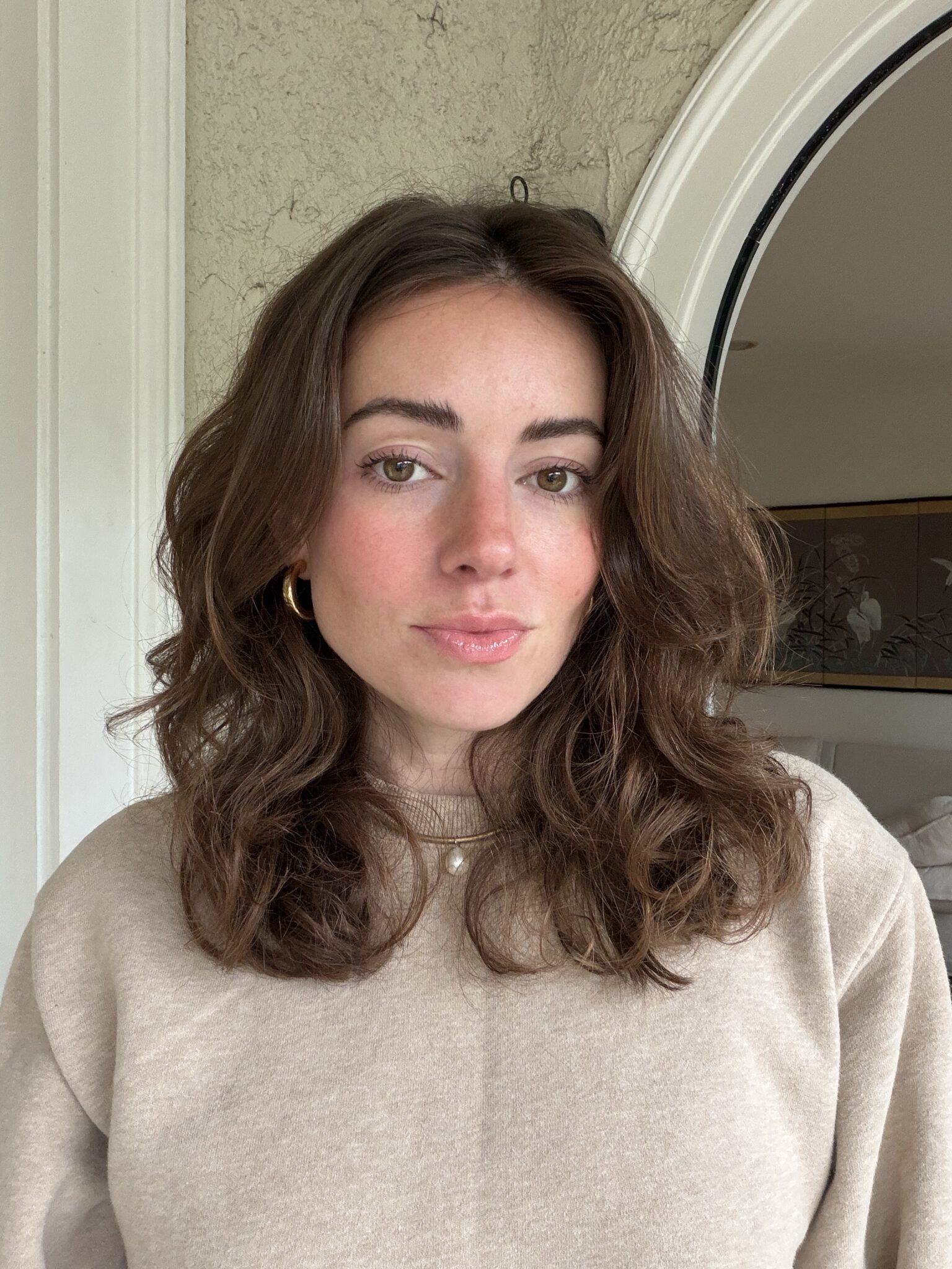 A woman with shoulder-length brown hair and a beige sweater stands indoors in front of a white wall and a round mirror.