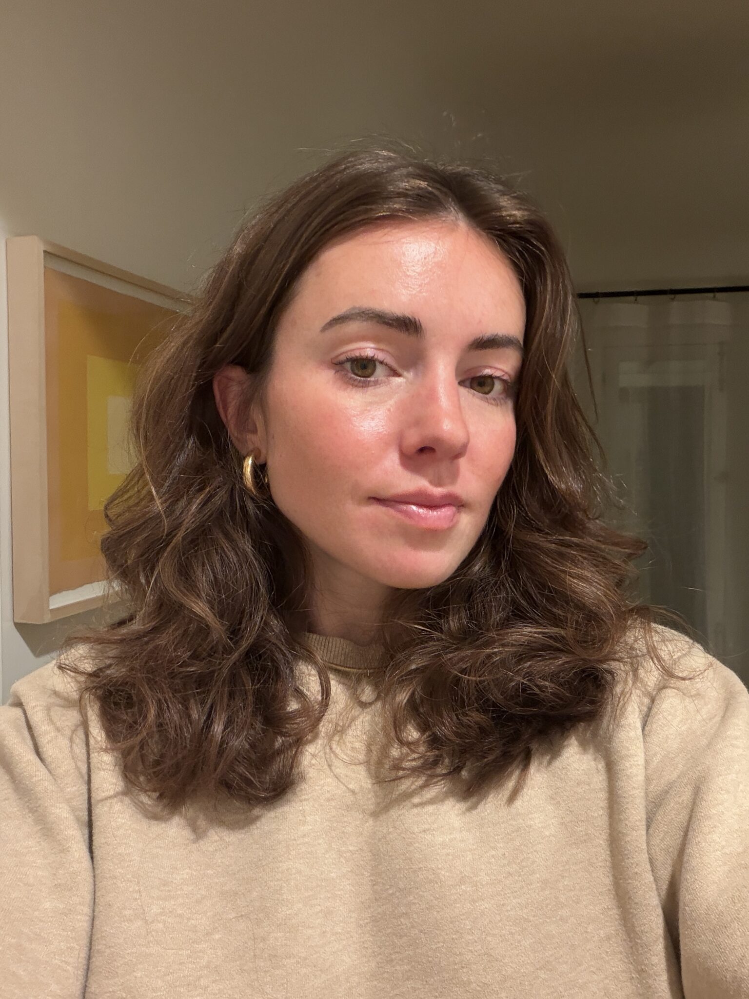 A woman with wavy brown hair and gold hoop earrings is wearing a beige sweater, posing indoors in front of a framed picture on the wall.