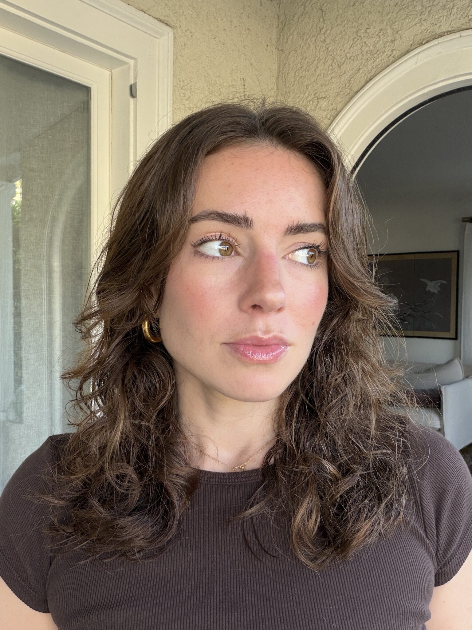 A woman with wavy brown hair and gold hoop earrings stands indoors near a window, looking slightly to her left. She is wearing a dark short-sleeved top.