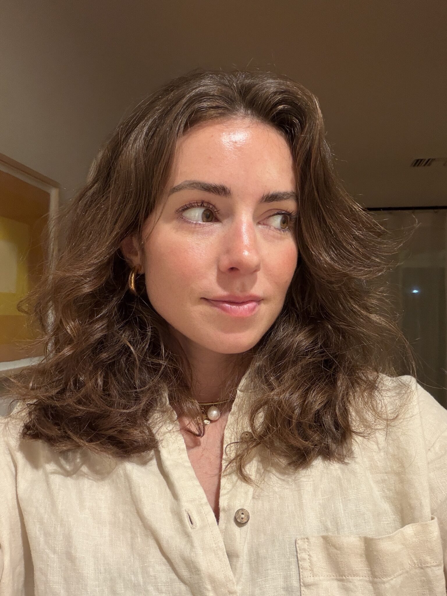 A woman with wavy brown hair and wearing a beige shirt looks to her left in a softly lit room.