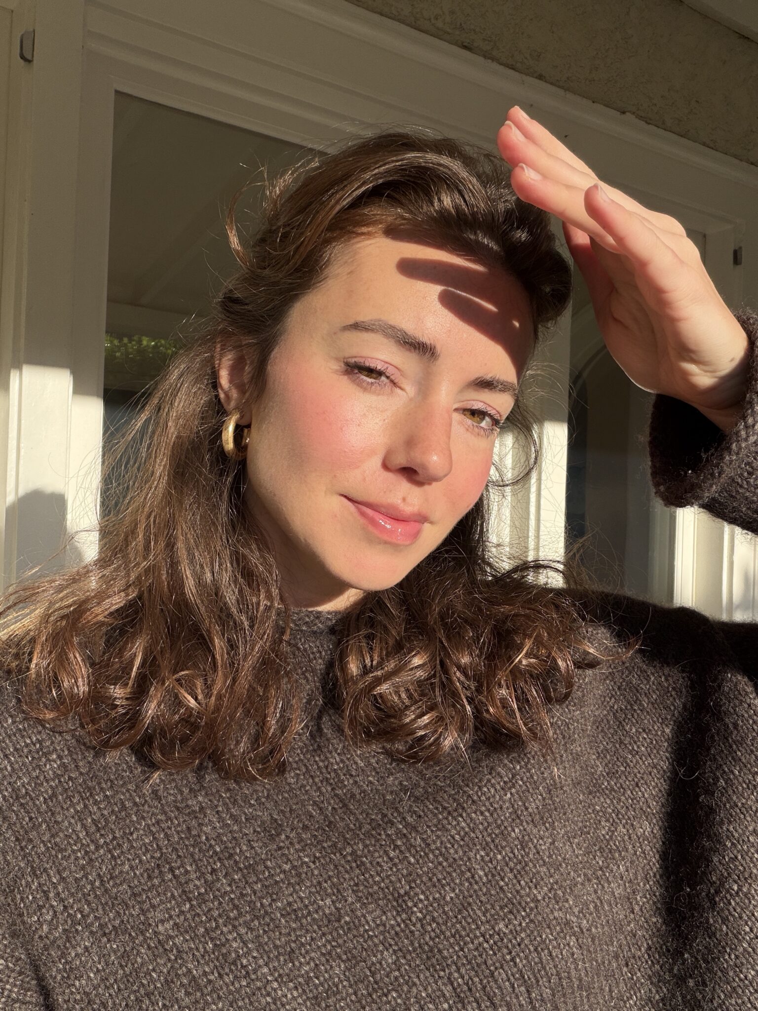 A woman with wavy brown hair shields her eyes from the sun with her hand while standing indoors near a window, wearing a brown sweater.