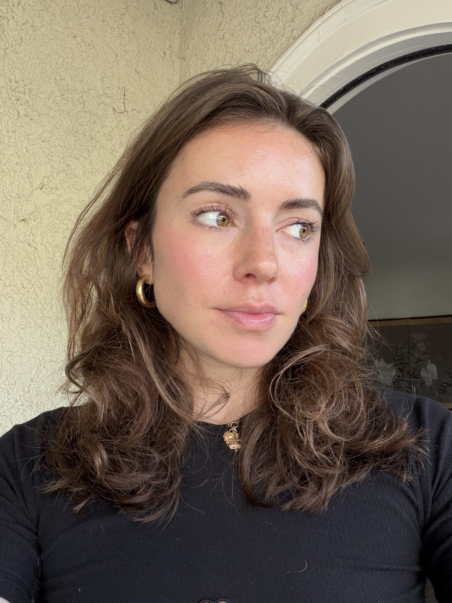 A woman with wavy brown hair, gold earrings, and a black top stands indoors looking to the side near a beige textured wall and an arched doorway.