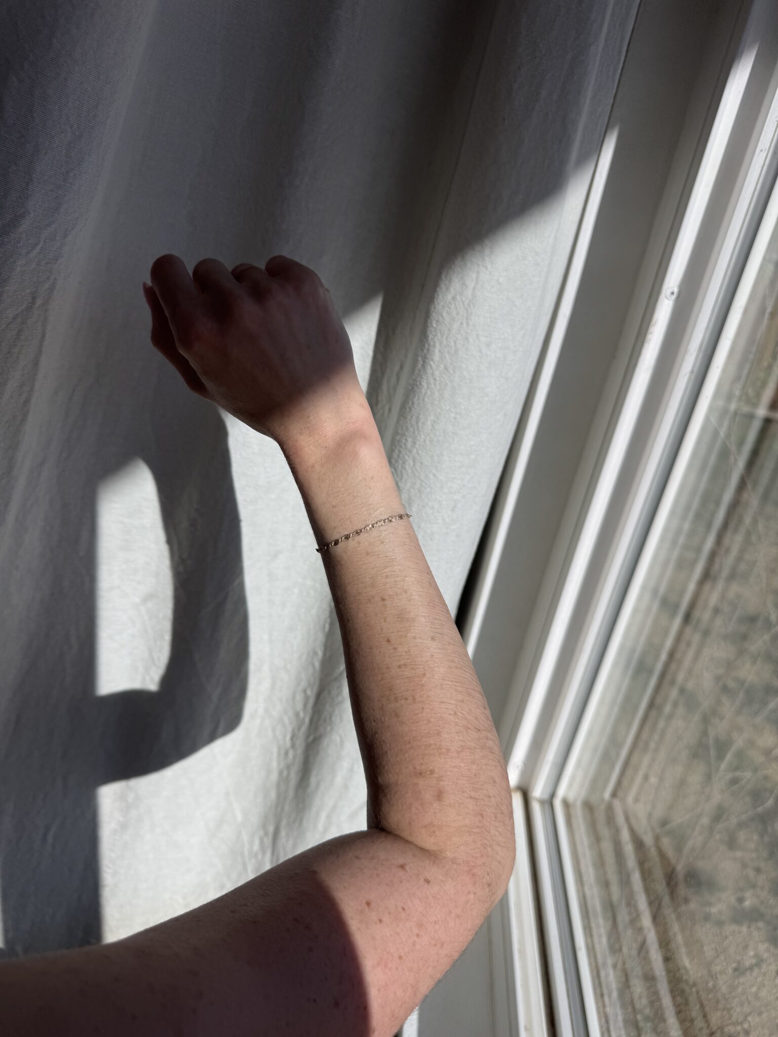 A person's arm with a delicate chain bracelet is raised near a sunlit window, casting a shadow on a white curtain.