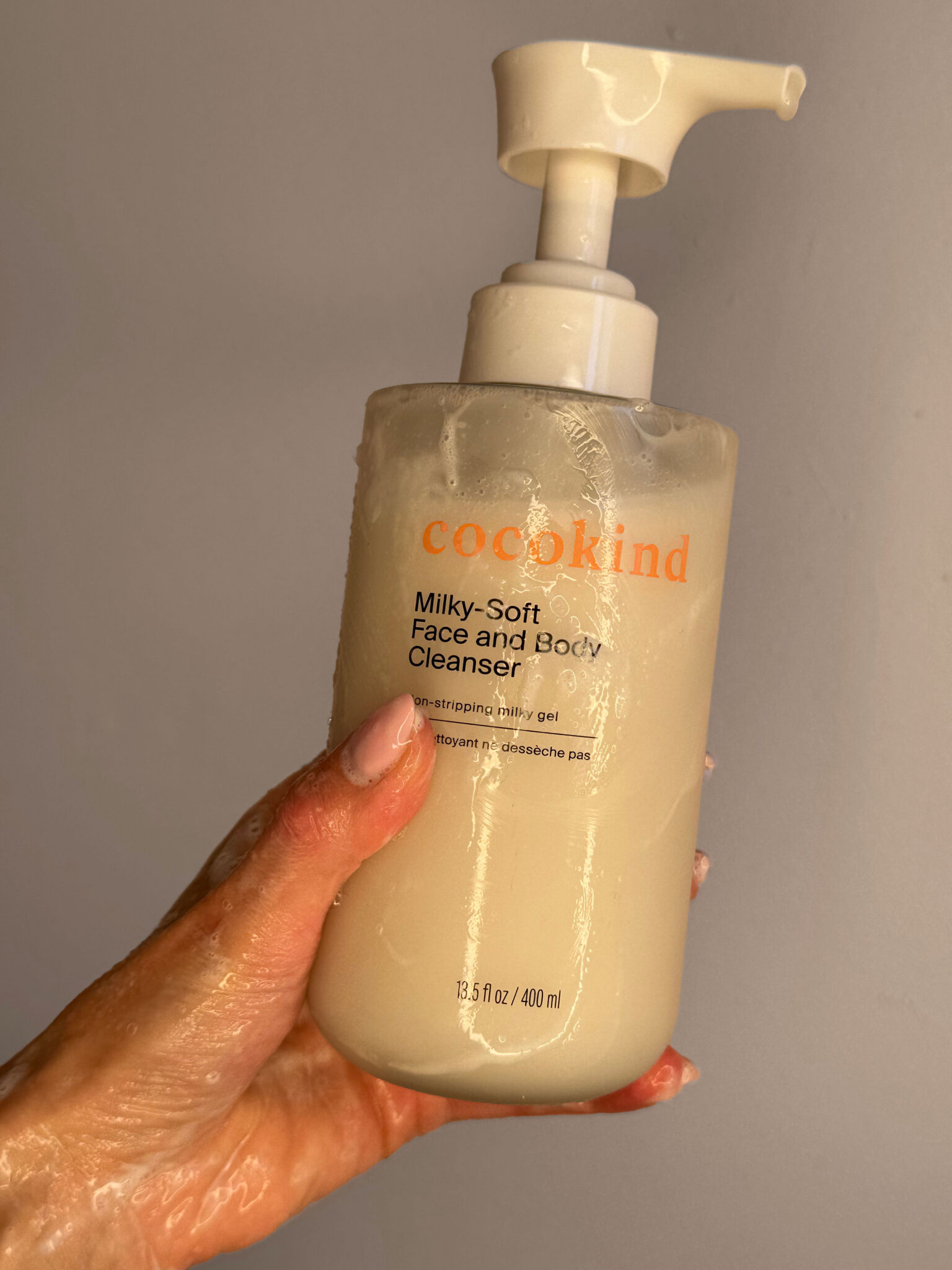 A hand holding a bottle of Cocokind Milky-Soft Face and Body Cleanser with a pump dispenser, partially covered in cleanser product.