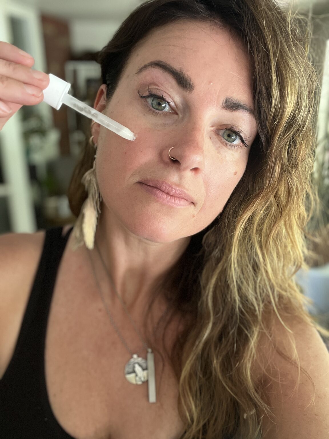 A woman with wavy hair applies facial serum to her cheek using a dropper. She wears a black top, feather earring, and two necklaces.