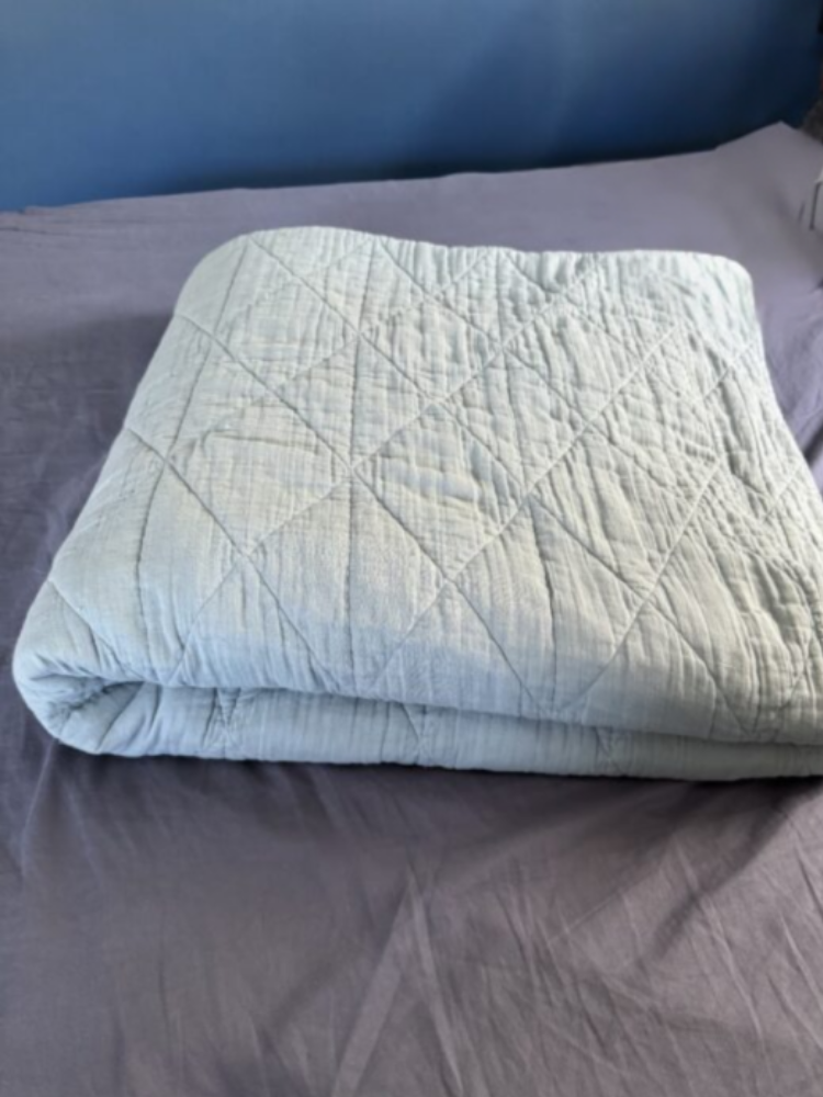 A light blue quilted blanket is neatly folded on a gray bedspread, with a blue wall in the background.