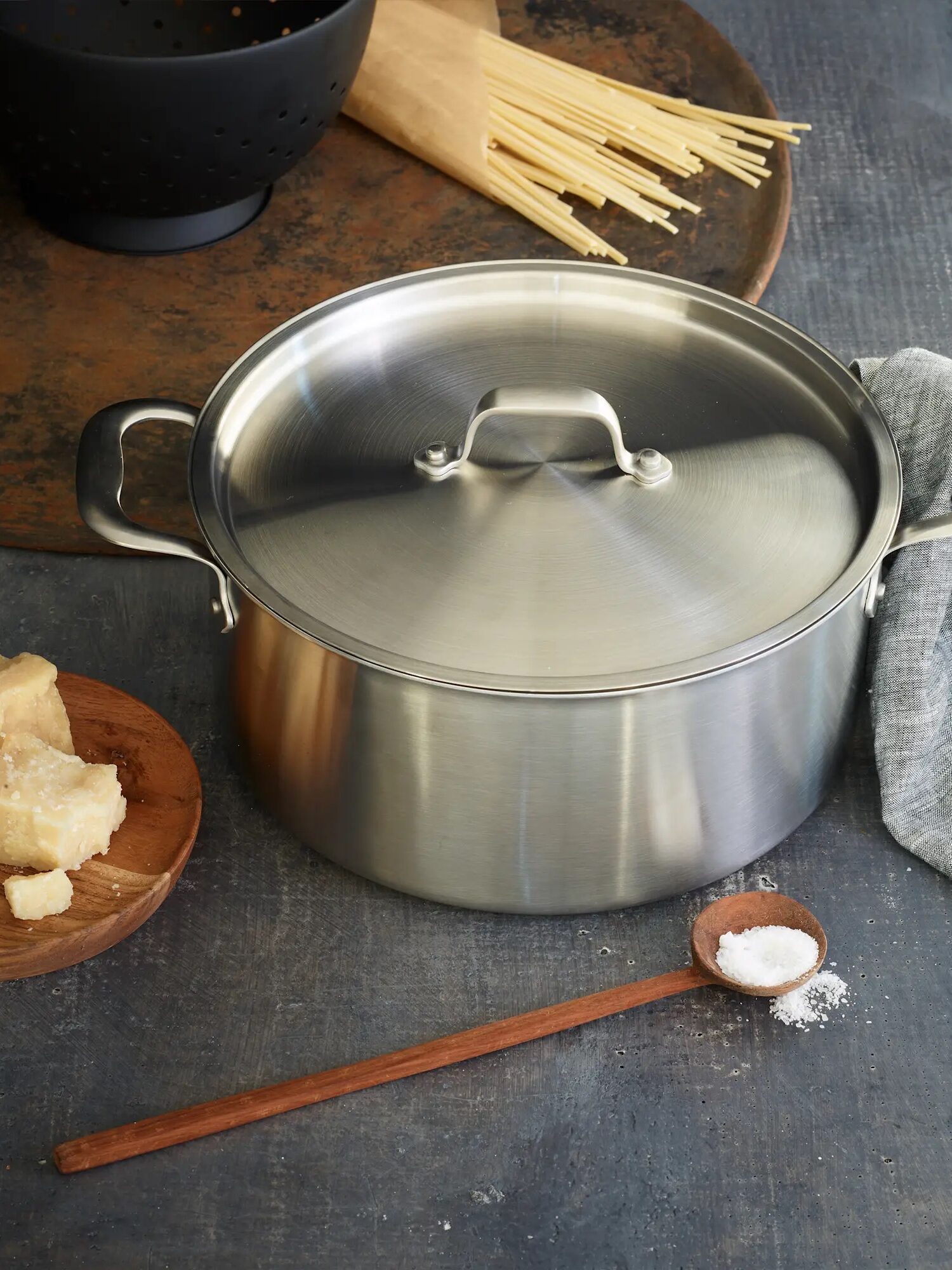 A stainless steel pot with a lid sits on a kitchen counter next to a plate of cheese, a wooden spoon with salt, uncooked spaghetti, and a gray cloth.