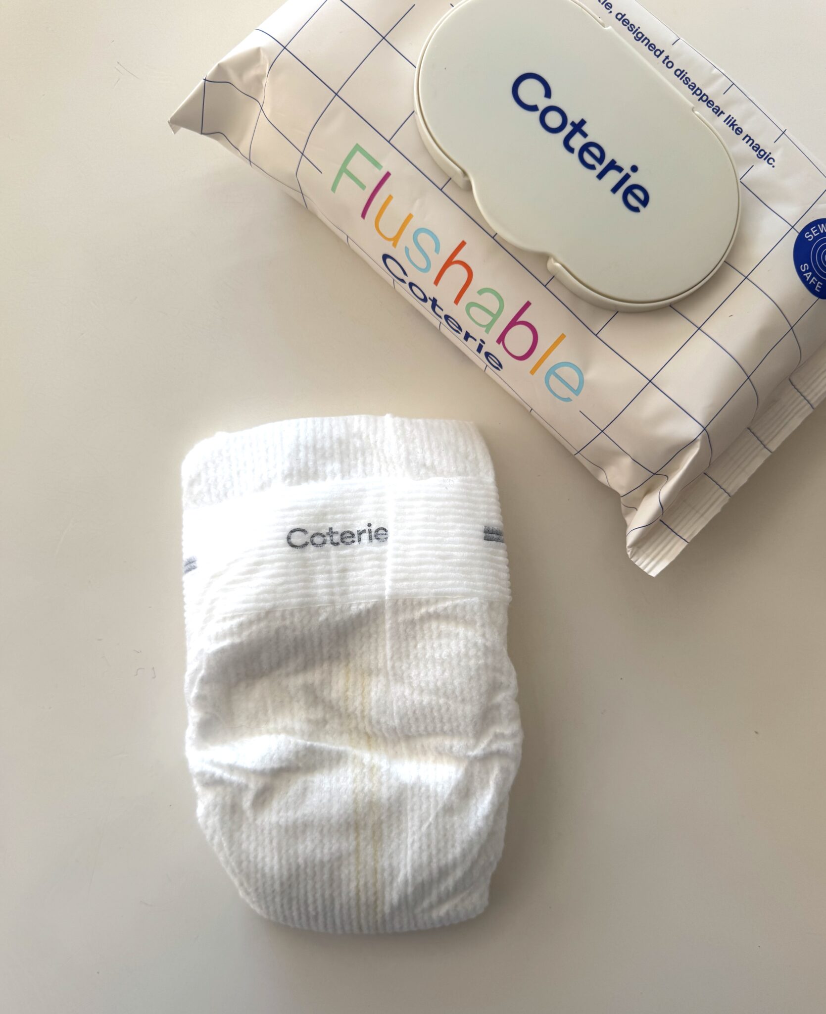 A Coterie brand diaper is placed on a white surface next to a pack of Coterie flushable wipes.