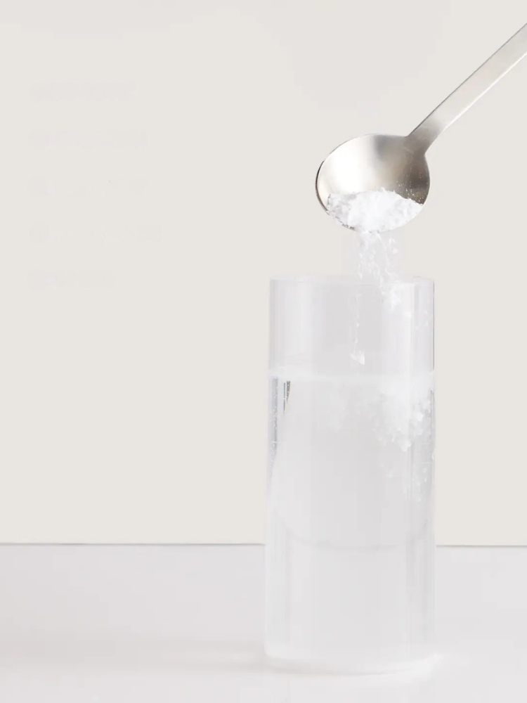 A spoon pours a white powder, likely sugar or salt, into a clear glass of water against a plain light background.