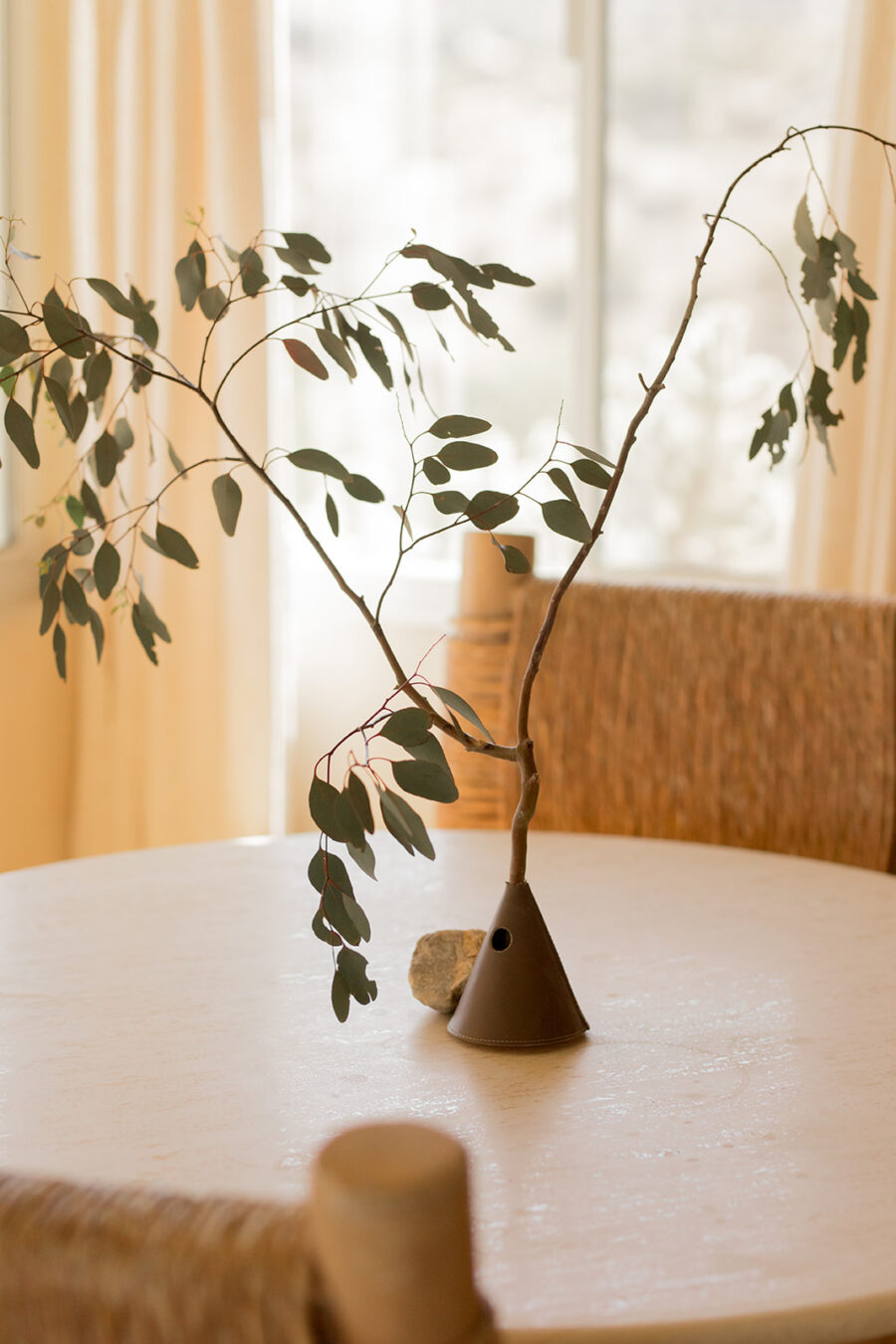 A slender branch with green leaves in a minimalist cone-shaped vase sits on a round table beside a small stone, evoking the calm of spiritual practice, with woven chairs and light curtains in the background.