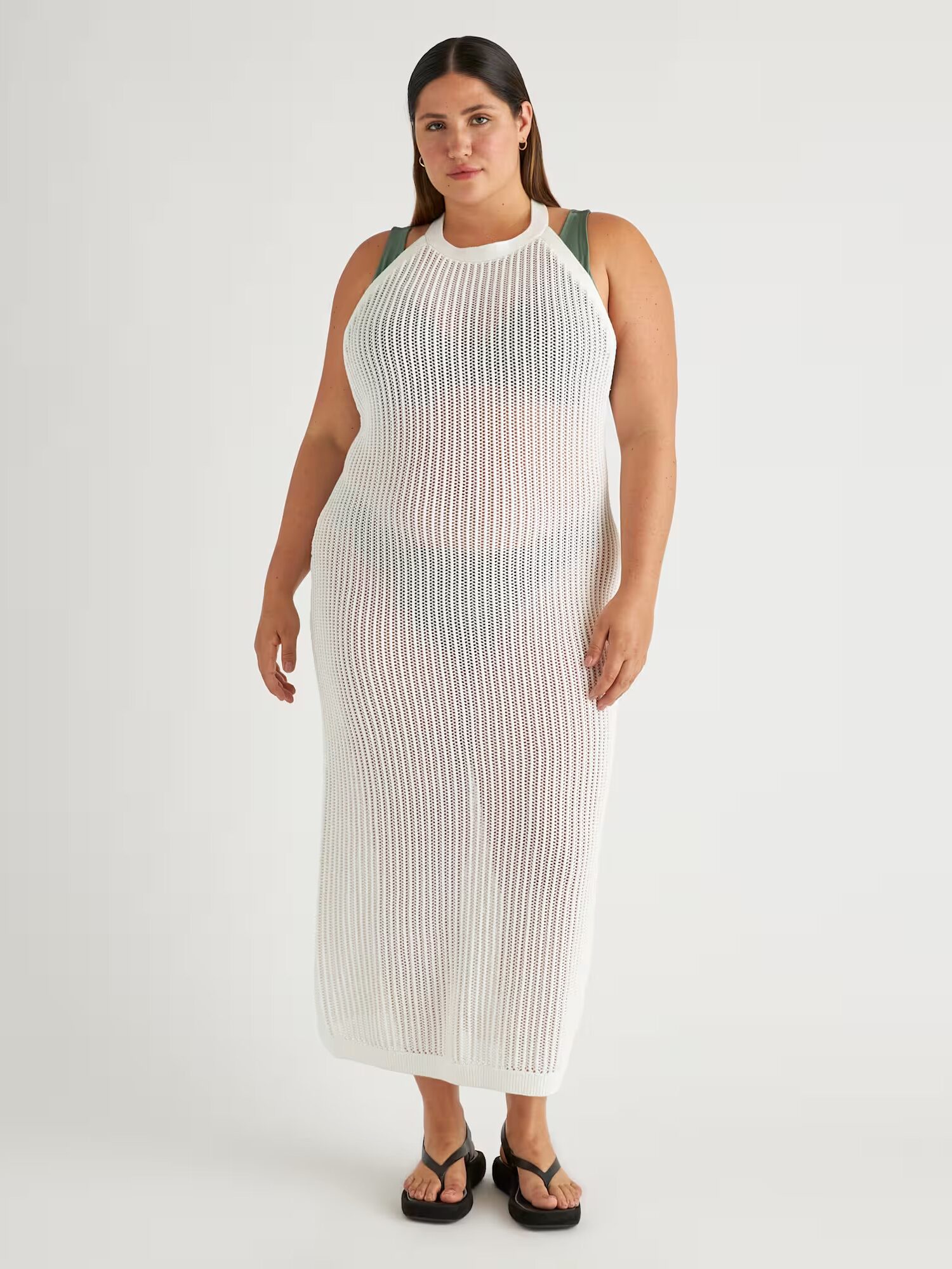 A woman stands facing forward wearing a sleeveless, long, white knit cover-up dress over a green bikini and black sandals against a plain background.