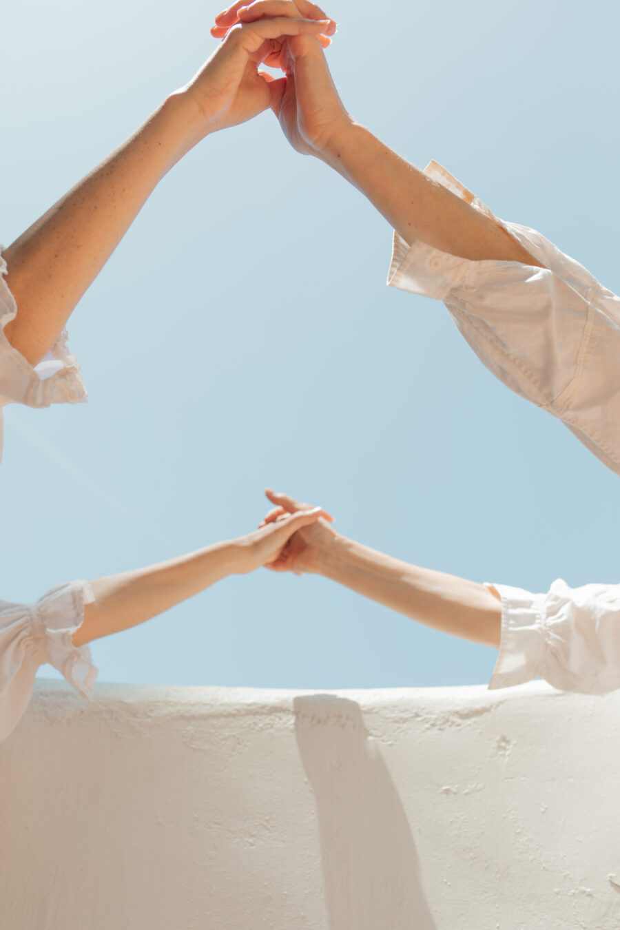 Two pairs of arms in white shirts reach up and touch hands against a bright blue sky and a white wall.